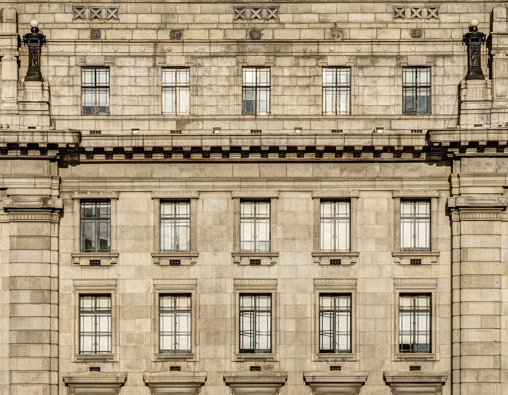 Monumental stone building facade featuring rows of glass windows, weathered masonry, classic architectural details